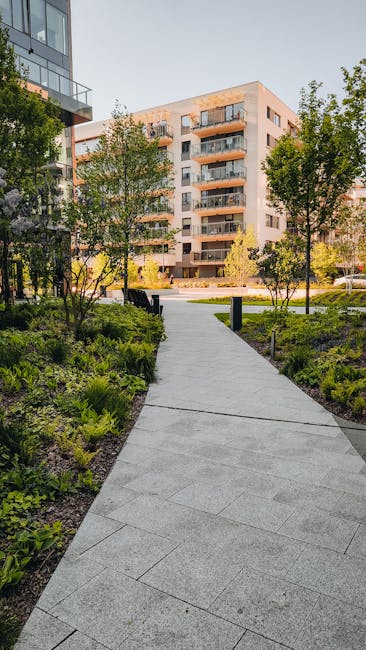A well-maintained urban garden pathway in White City, W12, leading towards a modern multi-storey residential building with balconies. The pathway is paved with light gray concrete slabs and curves gen